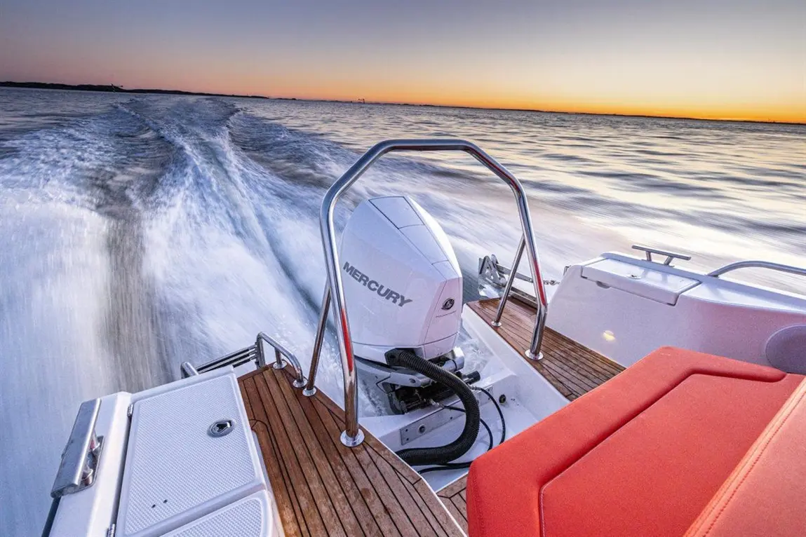 A boat with a Mercury engine speeds across calm water at sunset, showcasing a wooden deck and a vibrant sky.